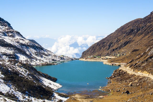 Turquoise Tsomgo Lake lying in a valley of brown, snow-streaked mountains with a road along its far shore
