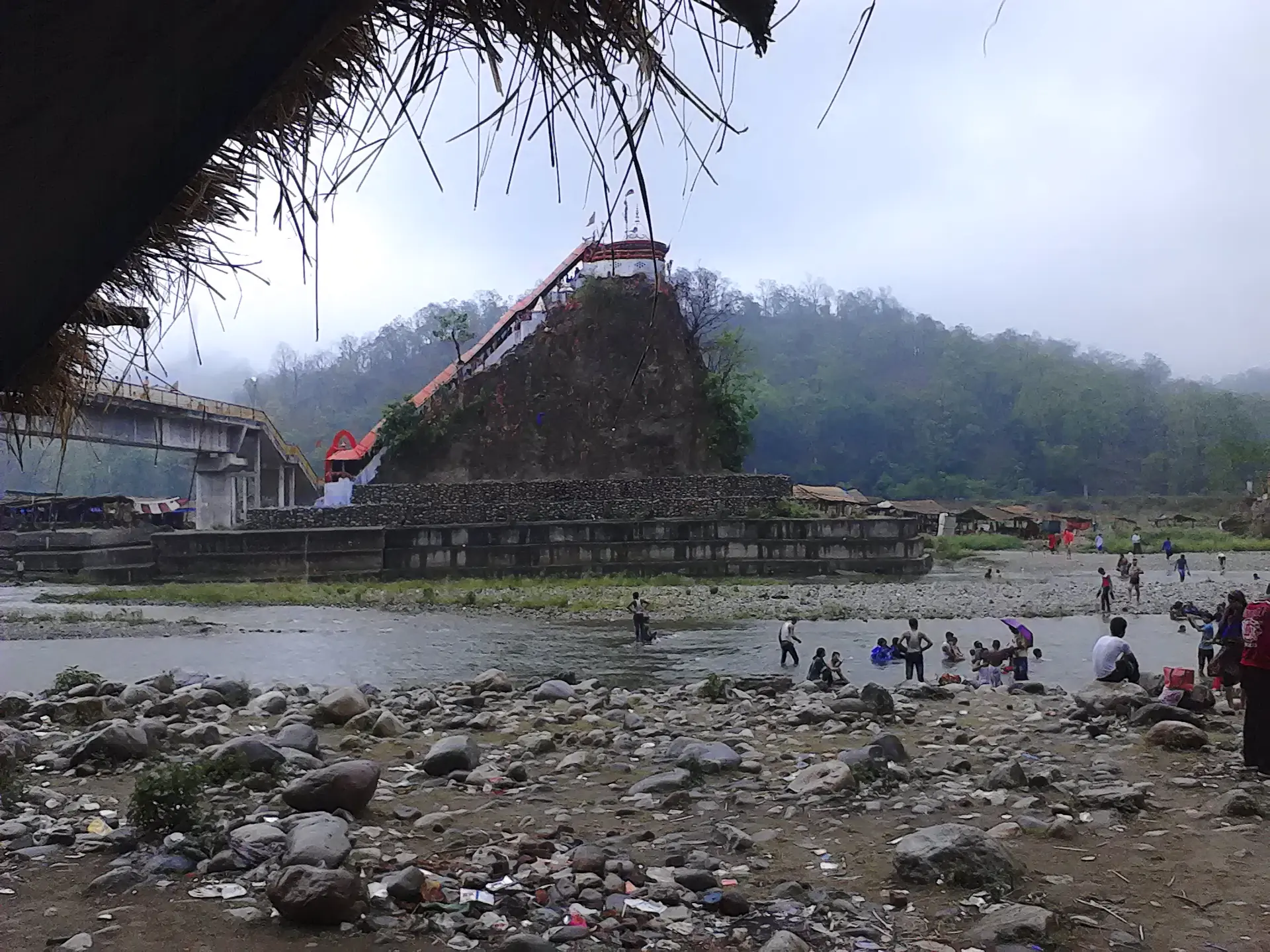 The red-roofed Garjiya Devi temple on a wooded rock in the Kosi river, with people on the stony riverbed below