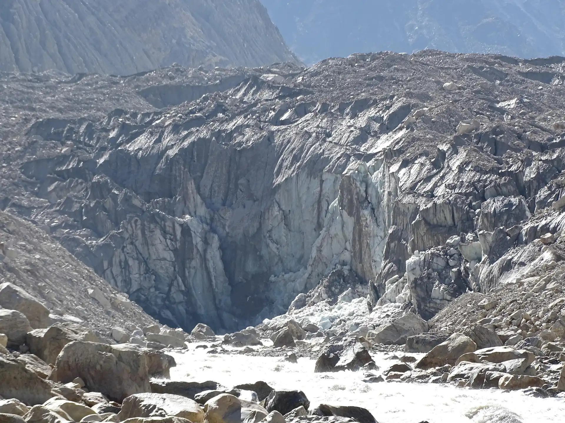 The grey broken ice cliff of the Gomukh snout with a meltwater stream and boulders in the foreground
