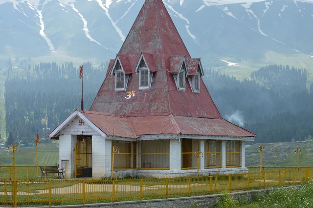 A red conical-roofed temple with white walls on a hilltop above a forested valley and snowy peaks