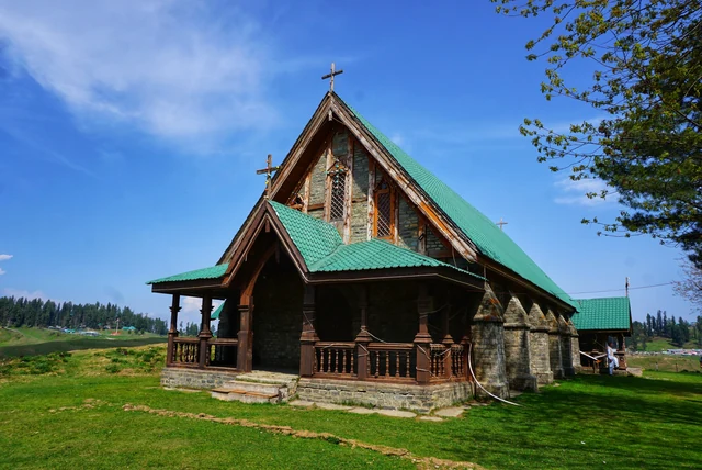 A grey stone church with a steep green roof and a wooden porch on a bright green lawn under a blue sky