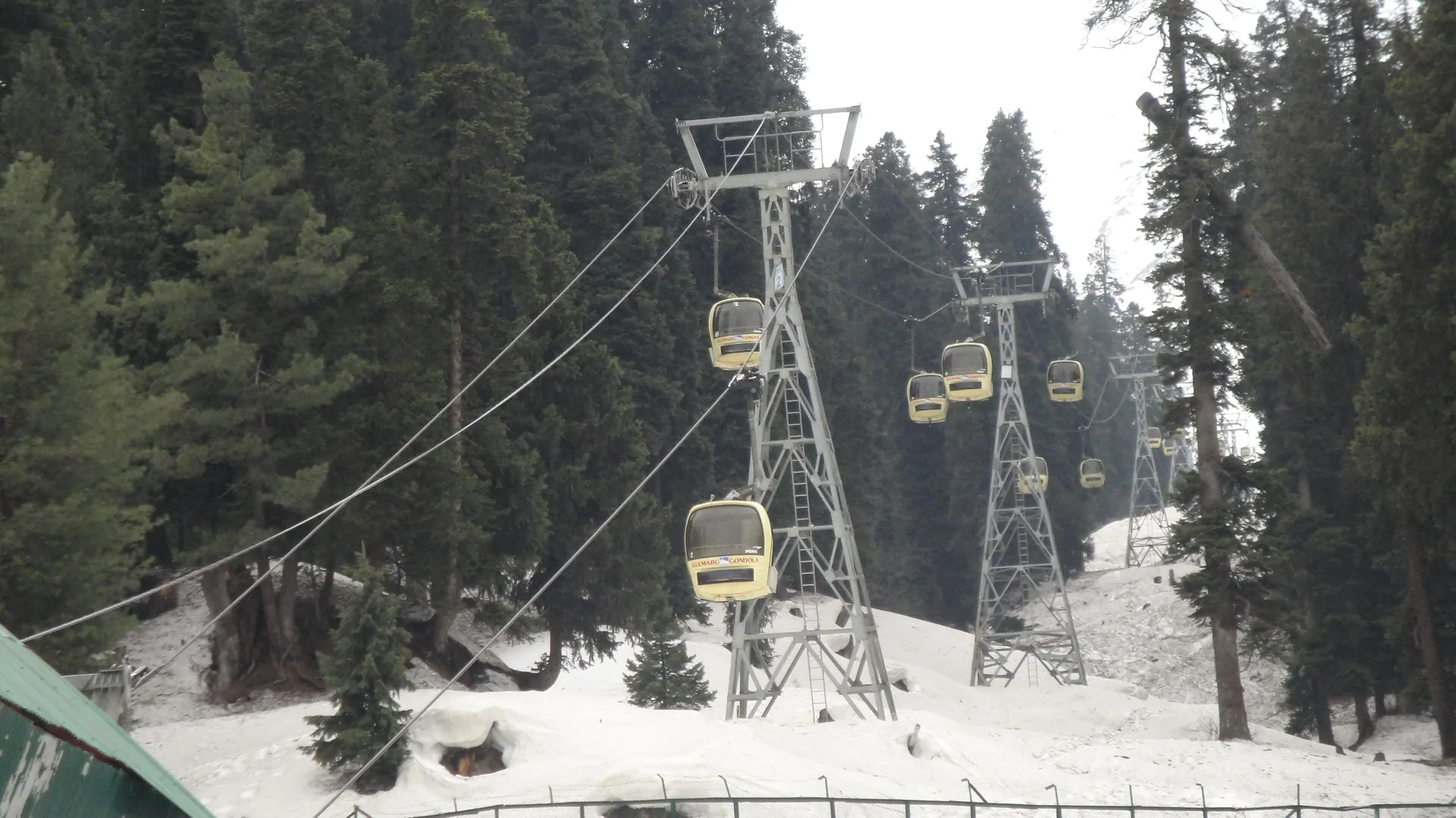 A steel tower carrying yellow gondola cabins on cables above snowy pine forest in Gulmarg