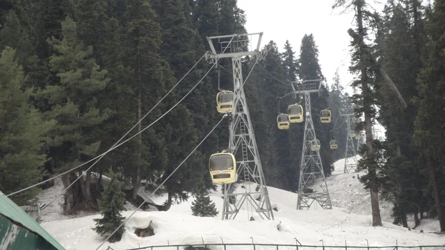 A steel tower carrying yellow gondola cabins on cables above snowy pine forest in Gulmarg