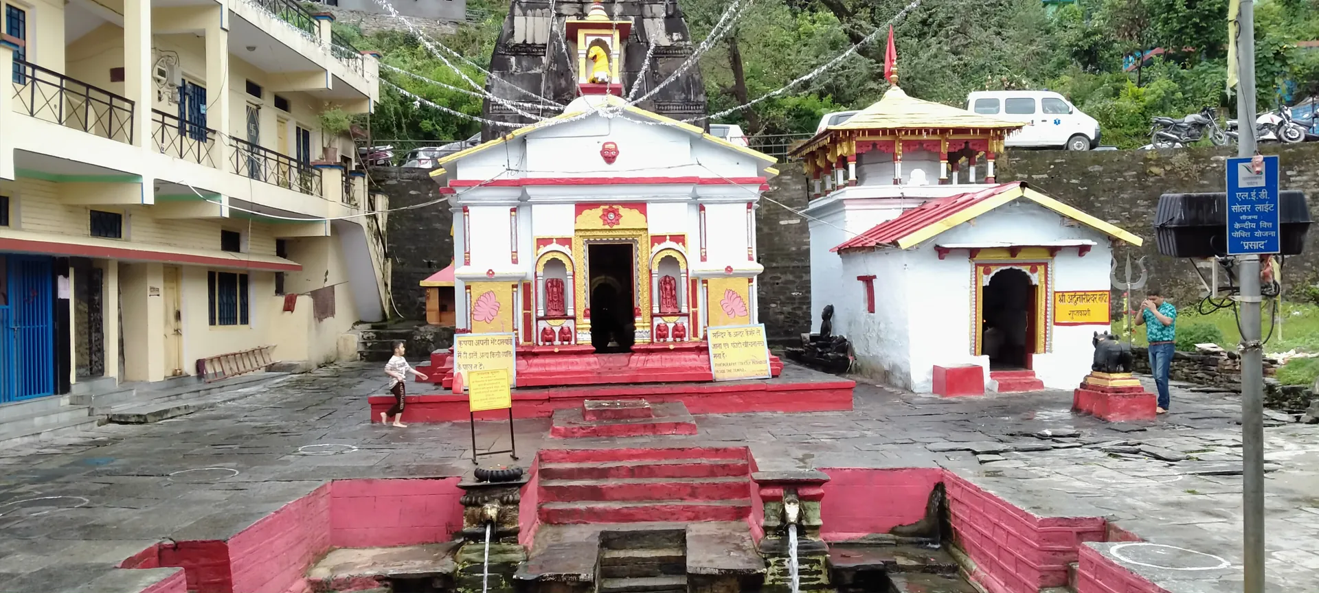 Two small shrines, one with a red-painted porch and a tower, beside a paved square in Guptakashi