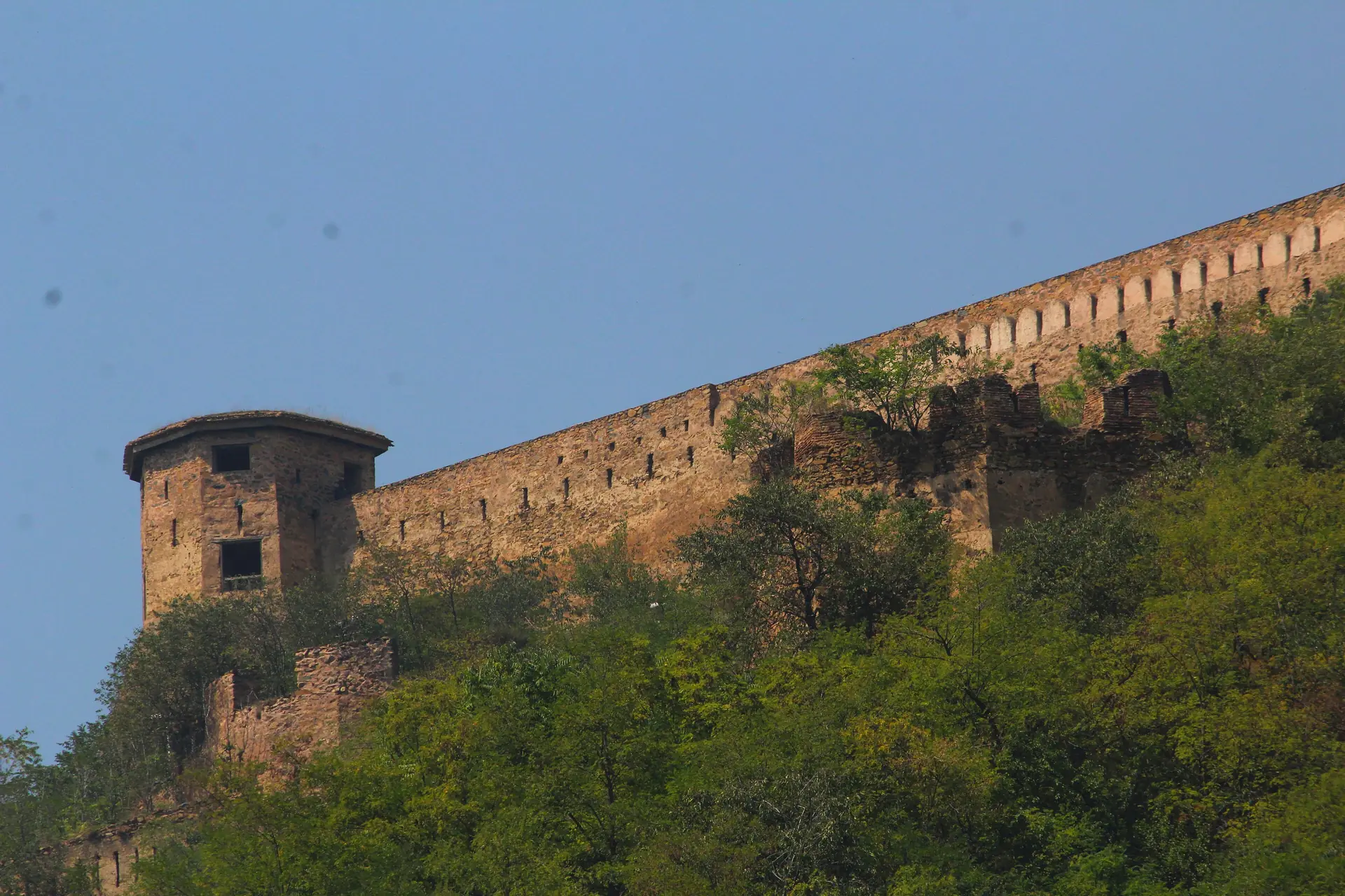 A long stone fort wall with a round tower along the green crest of Hari Parbat under a blue sky