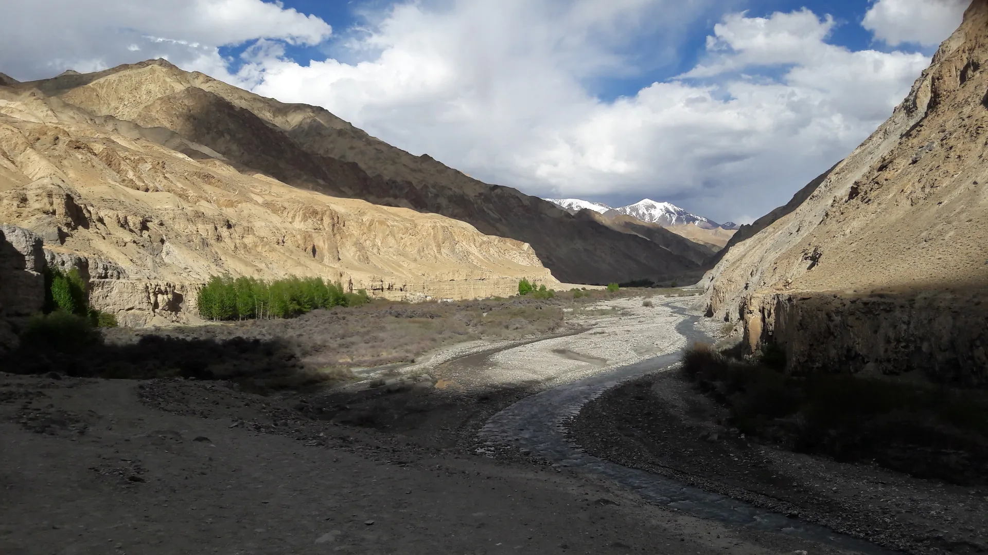 A wide gravel river bed winding between bare brown mountains and steep cliffs in the Markha valley under a cloudy sky
