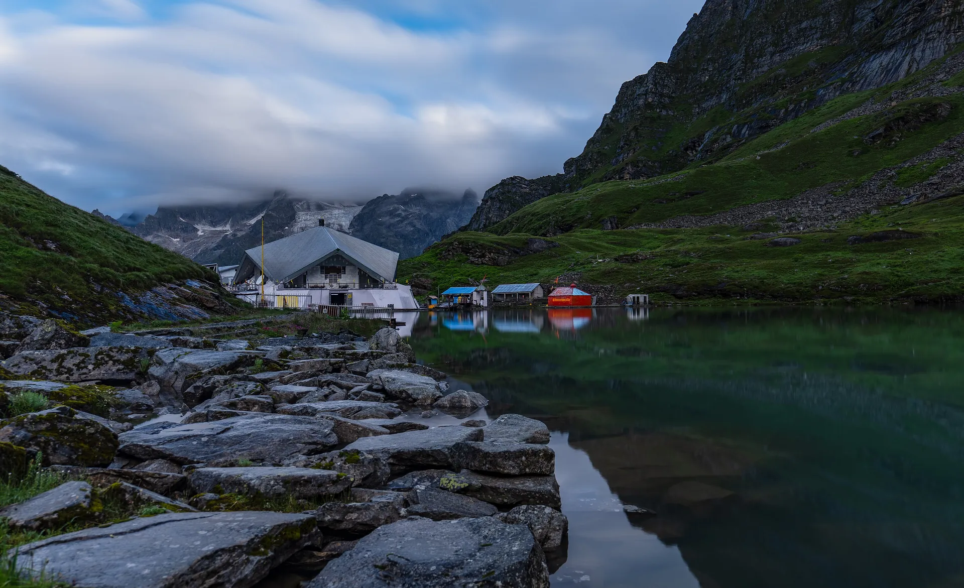 The white gurdwara of Hemkund Sahib beside its glacial lake, with rocky shore in front and mountains and cloud behind