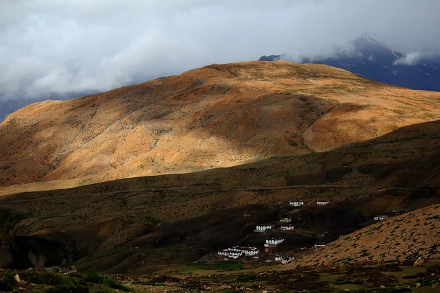 The white houses of Hikkim village on a bare high slope in Spiti, under a lit brown mountainside