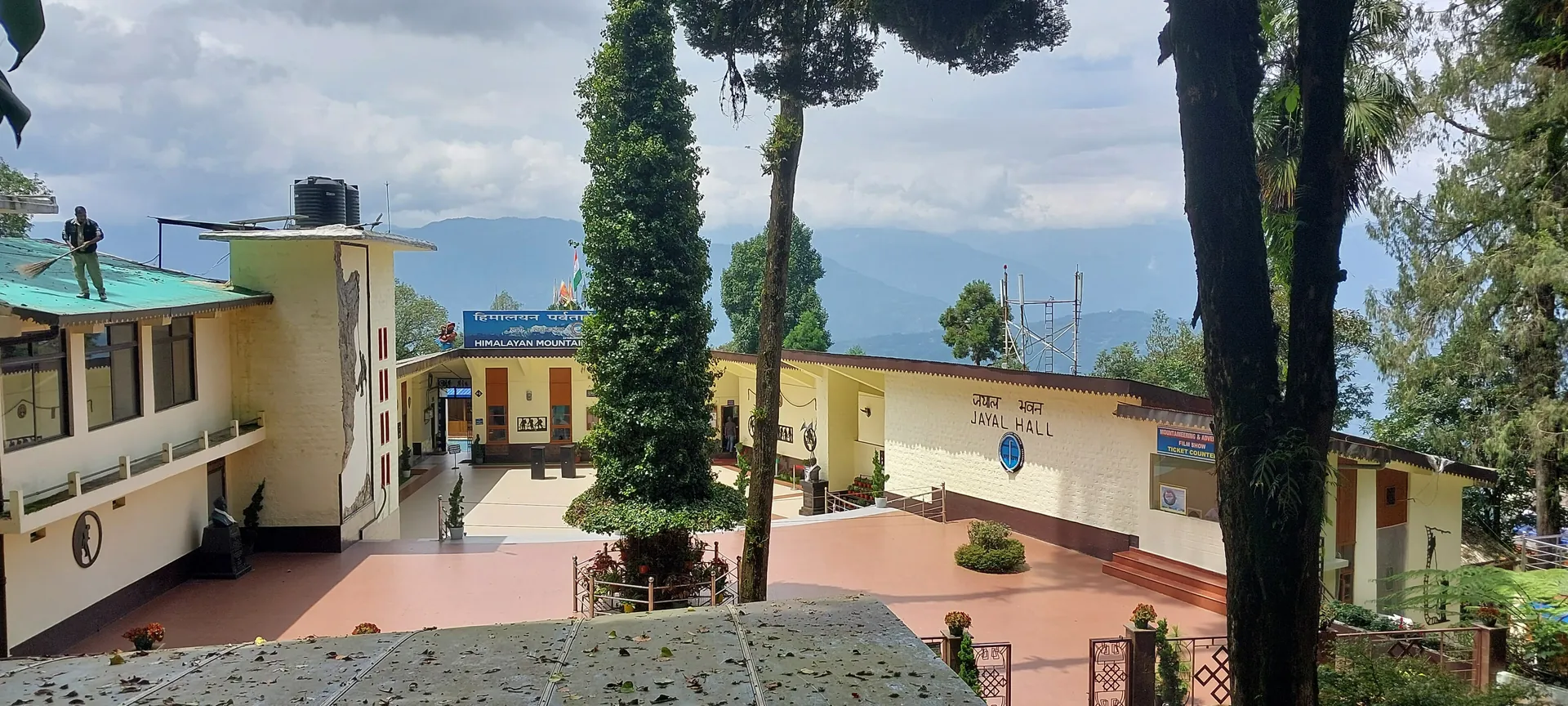 The pale yellow buildings of the Himalayan Mountaineering Institute around a paved courtyard with a tall tree and hills behind