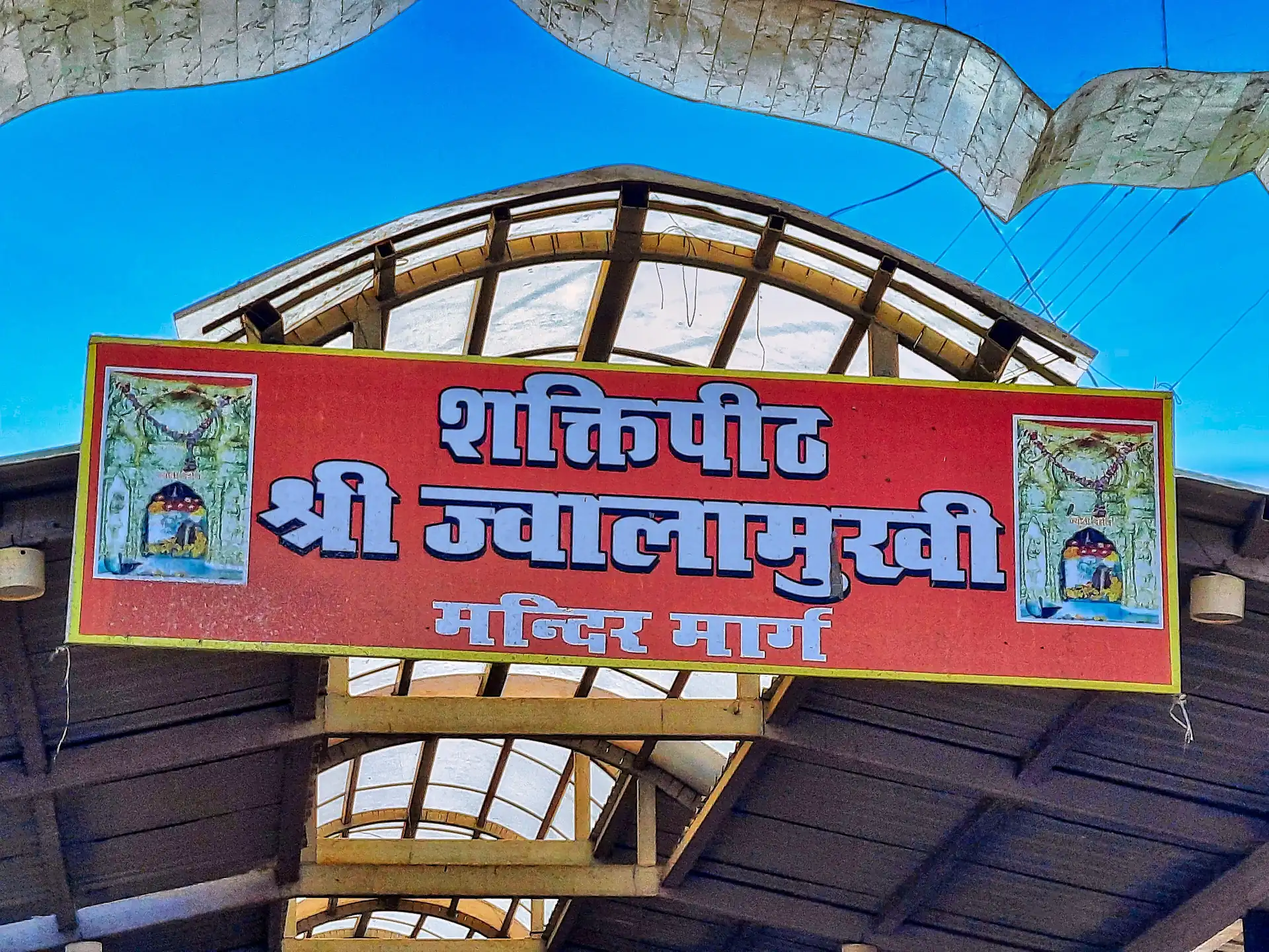 A red sign reading Shaktipeeth Shri Jwalamukhi Mandir Marg above a covered approach to the temple