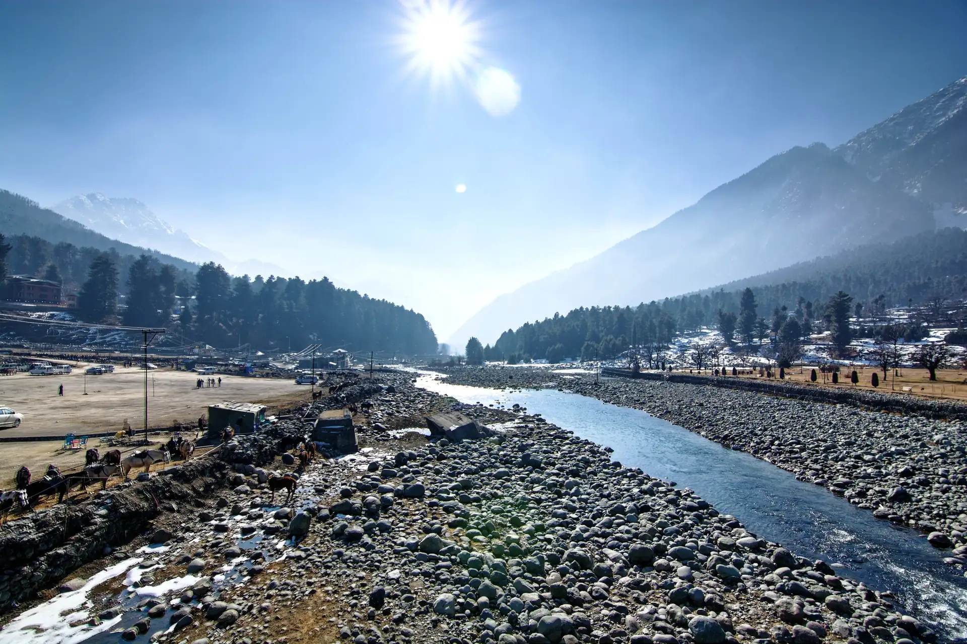 A pebble-banked stream in sunlight at Pahalgam with forested hills and a snow peak in the distance