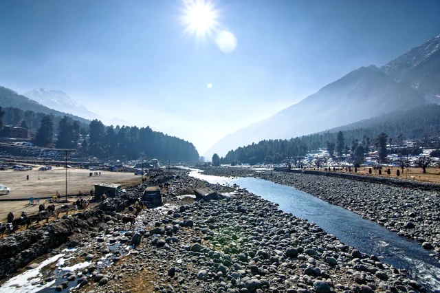 A pebble-banked stream in sunlight at Pahalgam with forested hills and a snow peak in the distance