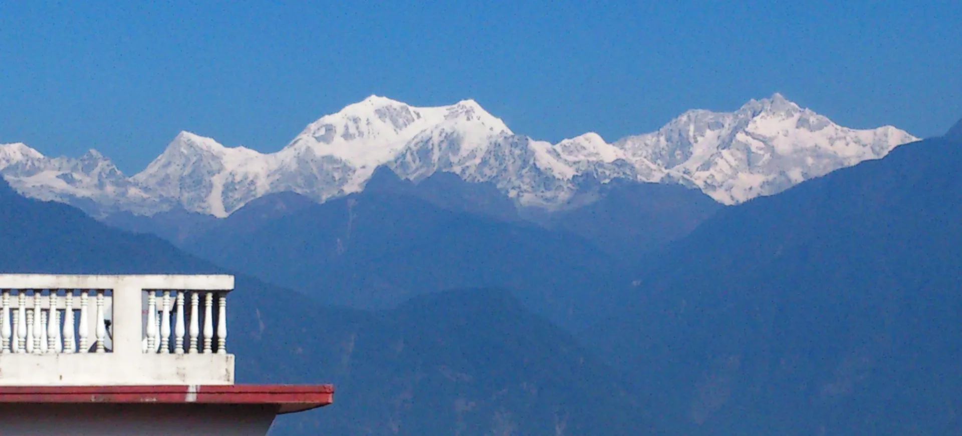 A snow-covered Kanchenjunga range under blue sky above forested ridges, with a white balcony rail in the foreground