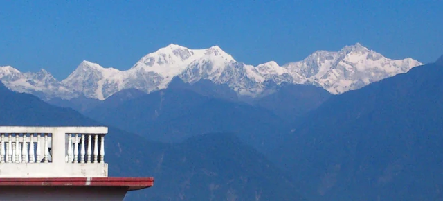 A snow-covered Kanchenjunga range under blue sky above forested ridges, with a white balcony rail in the foreground