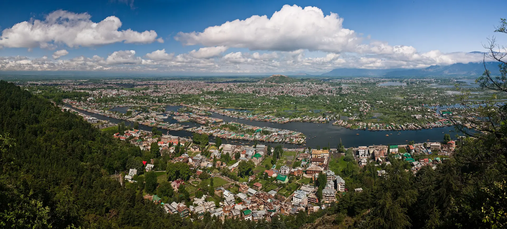 A wide aerial view of Srinagar's rooftops, the Jhelum river and Dal Lake under a blue sky with cloud