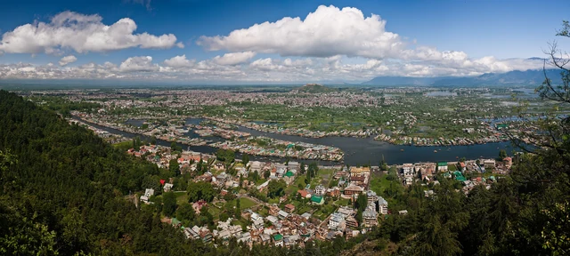 A wide aerial view of Srinagar's rooftops, the Jhelum river and Dal Lake under a blue sky with cloud