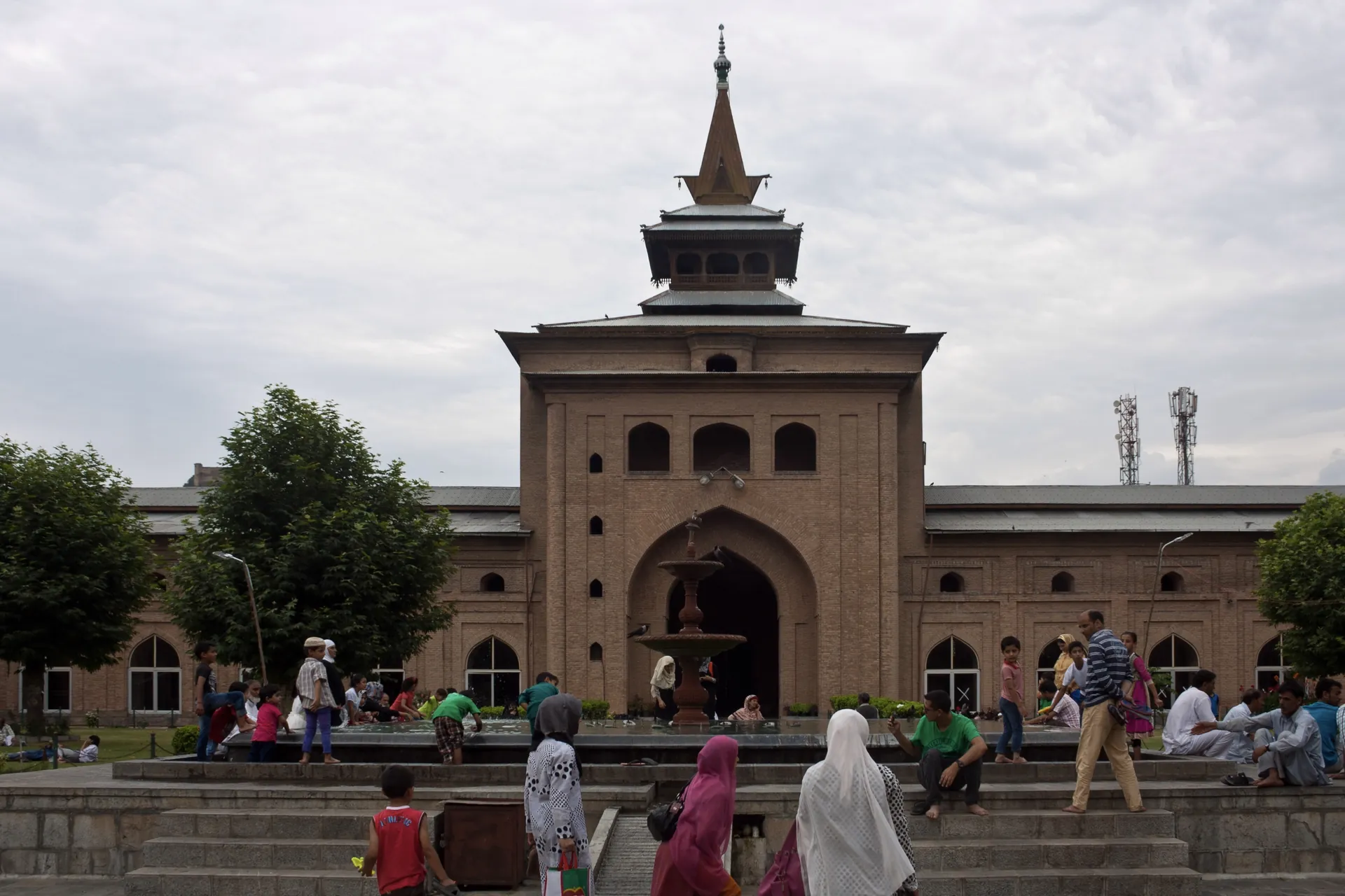 The brick facade of the Jamia Masjid with a pyramidal gateway and spire above a courtyard with visitors