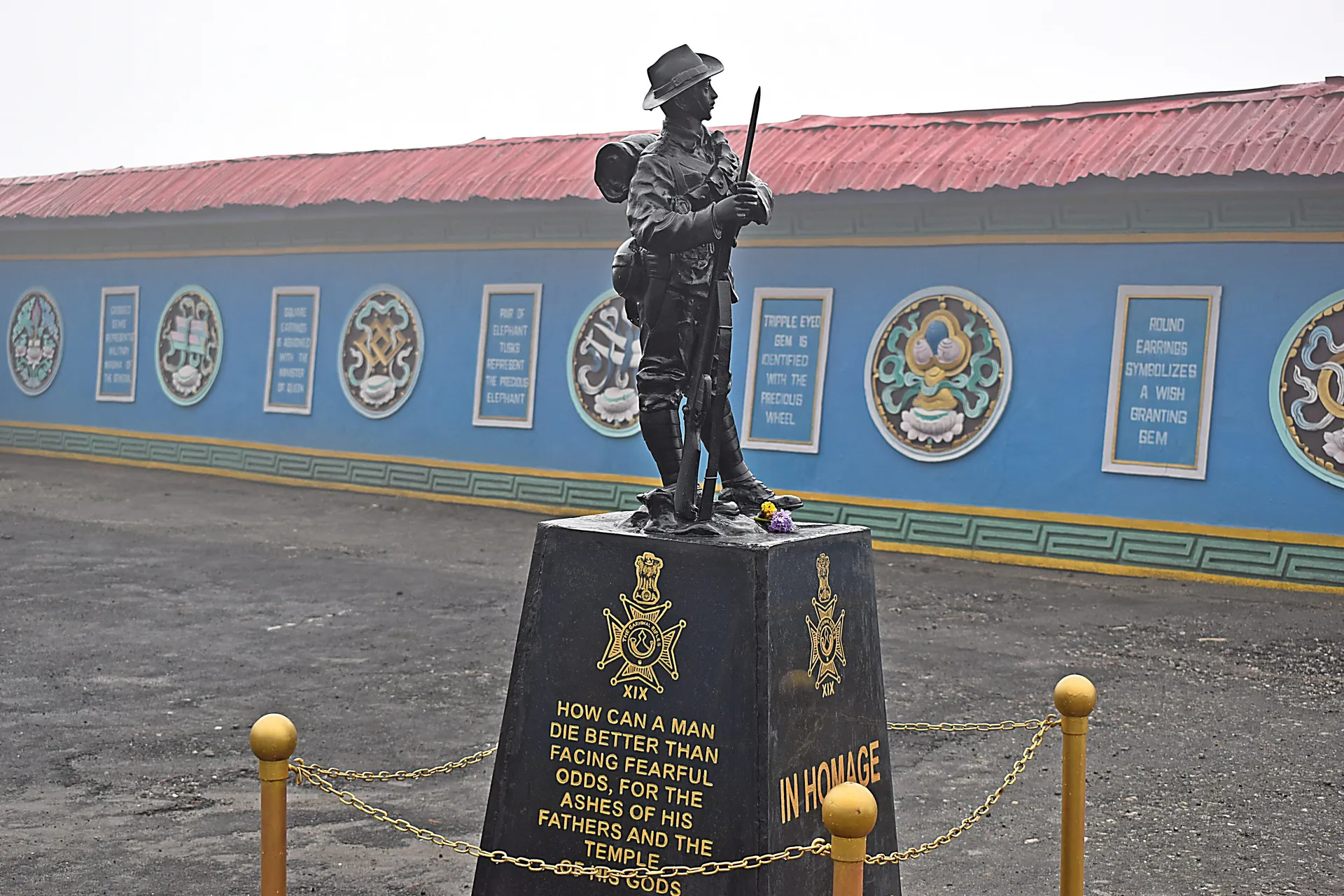A bronze statue of a soldier with a rifle on a black plinth in front of a long blue memorial wall with painted emblems
