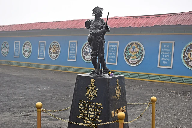 A bronze statue of a soldier with a rifle on a black plinth in front of a long blue memorial wall with painted emblems