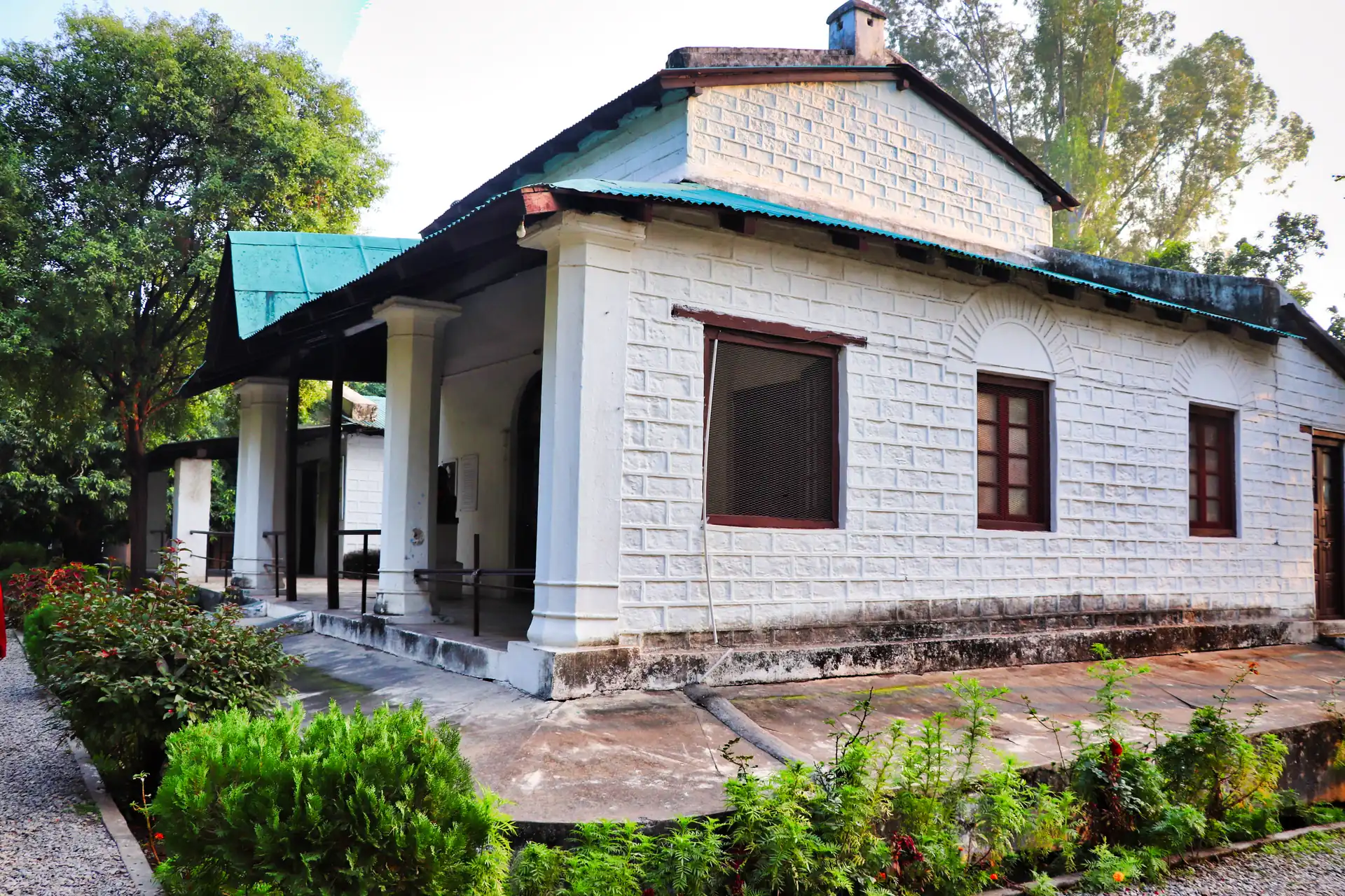 A white stone bungalow with a green metal roof and a small verandah among garden trees