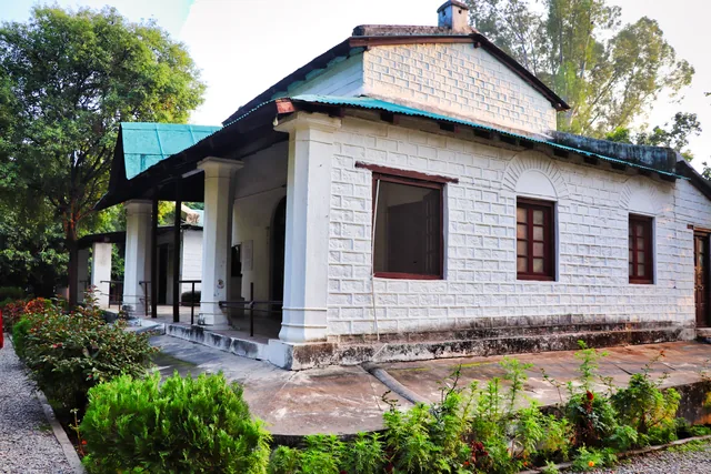 A white stone bungalow with a green metal roof and a small verandah among garden trees