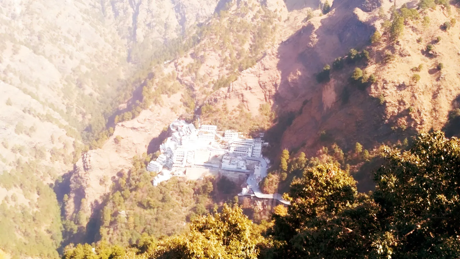 The Vaishno Devi shrine buildings set into a fold of the Trikuta mountain, seen from far above in soft light