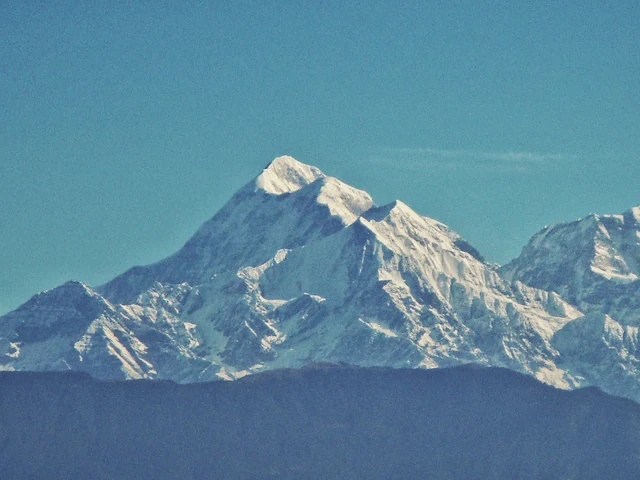 The snow-covered pyramid of Trishul peak against a clear blue sky above dark foothills