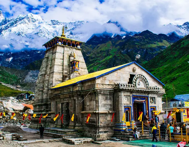 The grey stone Kedarnath Temple with its tall tower and a yellow roof, below green slopes and snow-covered peaks