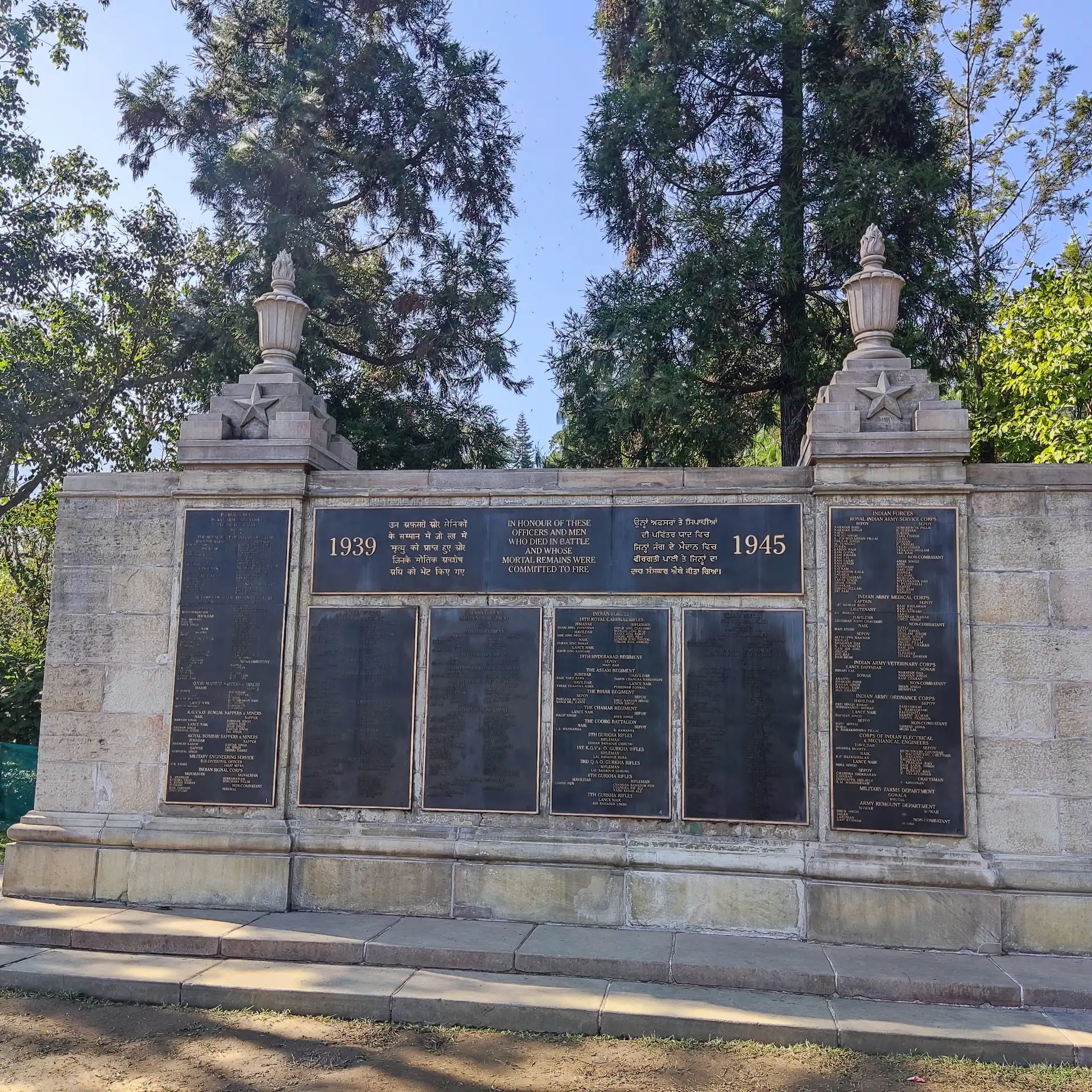 The stone memorial wall of the Kohima War Cemetery with dark inscribed panels between two pillars, under tall trees