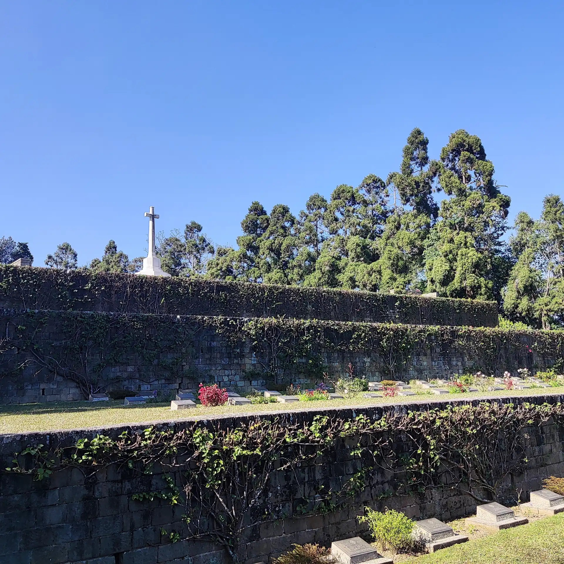 Terraced green lawns and stone walls of the Kohima War Cemetery rising to a white cross against a blue sky