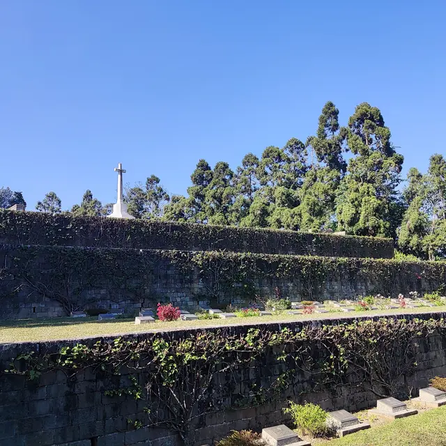 Terraced green lawns and stone walls of the Kohima War Cemetery rising to a white cross against a blue sky