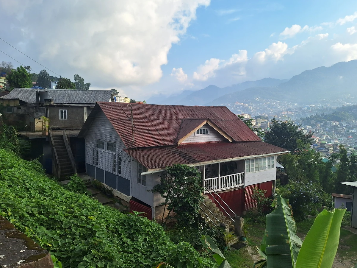 Wooden houses with red tin roofs on a green slope in Kohima Village, with the city and blue hills behind under white clouds