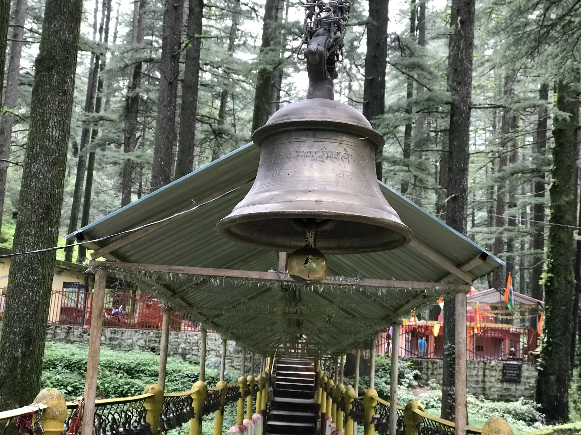 A large bronze bell hanging under a small green roof at the top of steps among tall deodar trees at Tarkeshwar Mahadev
