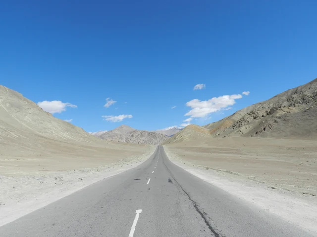 A paved highway running between bare brown and grey mountain slopes under a blue sky with scattered clouds