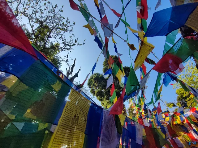 A covered walkway under a canopy of colourful prayer flags beside a red prayer wheel rack and a stone bench at the Mahakal temple