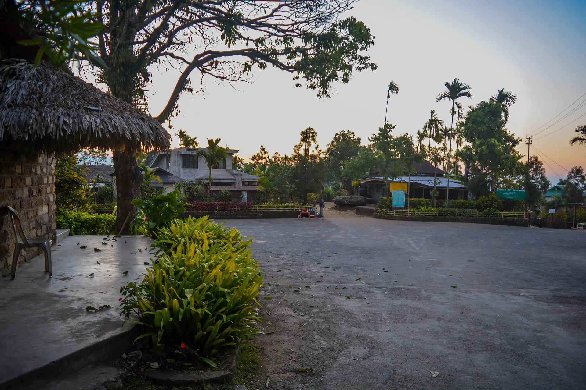A quiet paved village square at dusk with a thatched shelter, palms and low houses lit by the low sun