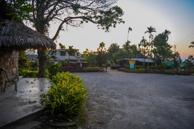 A quiet paved village square at dusk with a thatched shelter, palms and low houses lit by the low sun