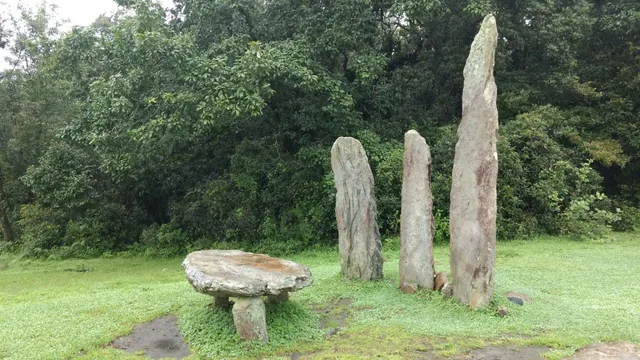 Three tall standing stones and a flat table stone on a green lawn in front of the dark forest of the Mawphlang sacred grove