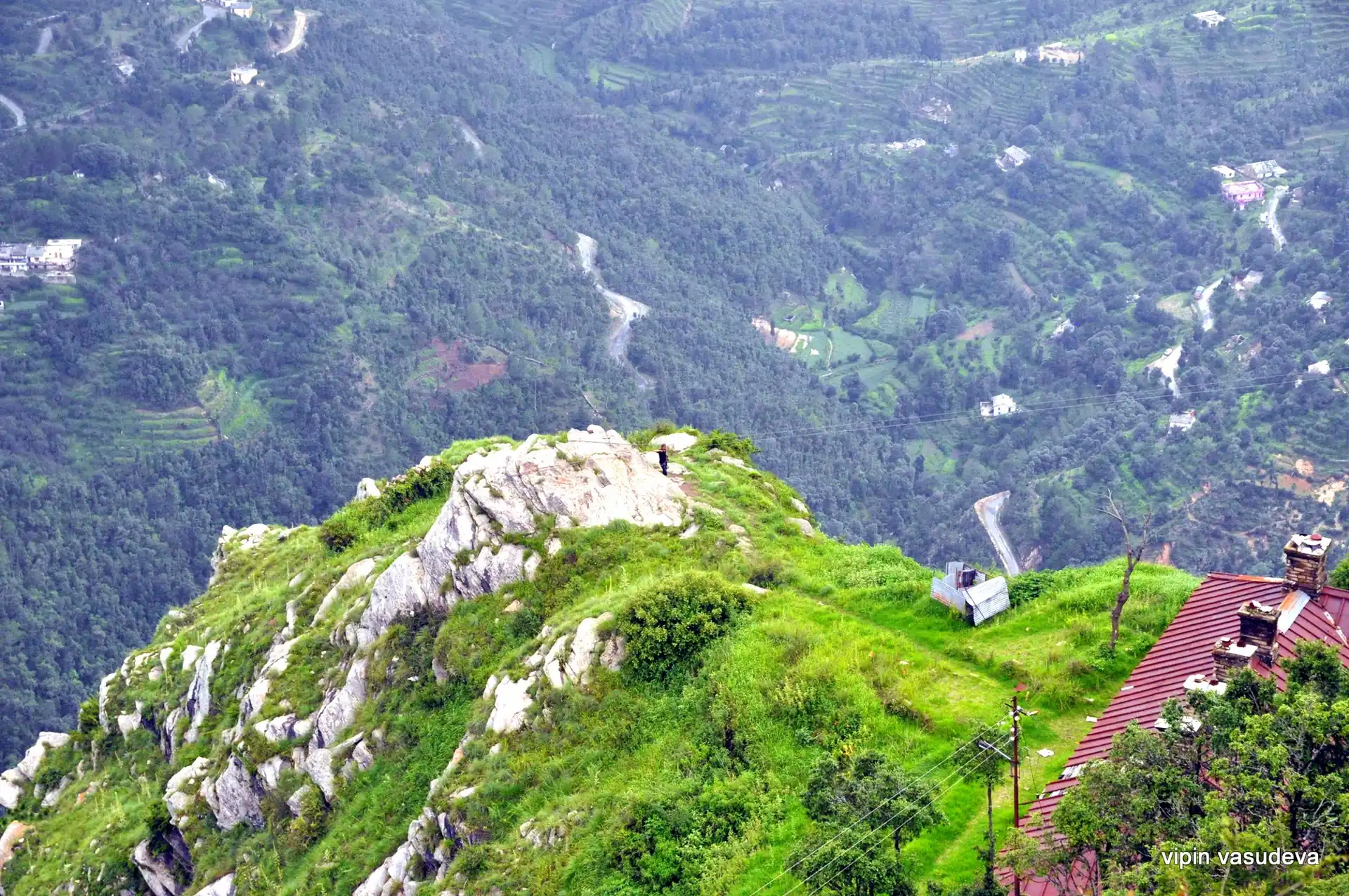 A grassy rock-topped ridge above terraced green slopes and a winding road in the valley below Mukteshwar