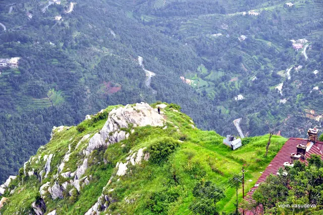 A grassy rock-topped ridge above terraced green slopes and a winding road in the valley below Mukteshwar