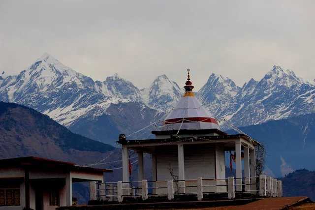 A small white temple with a red-banded dome on a ridge facing a line of snow peaks near Munsiyari