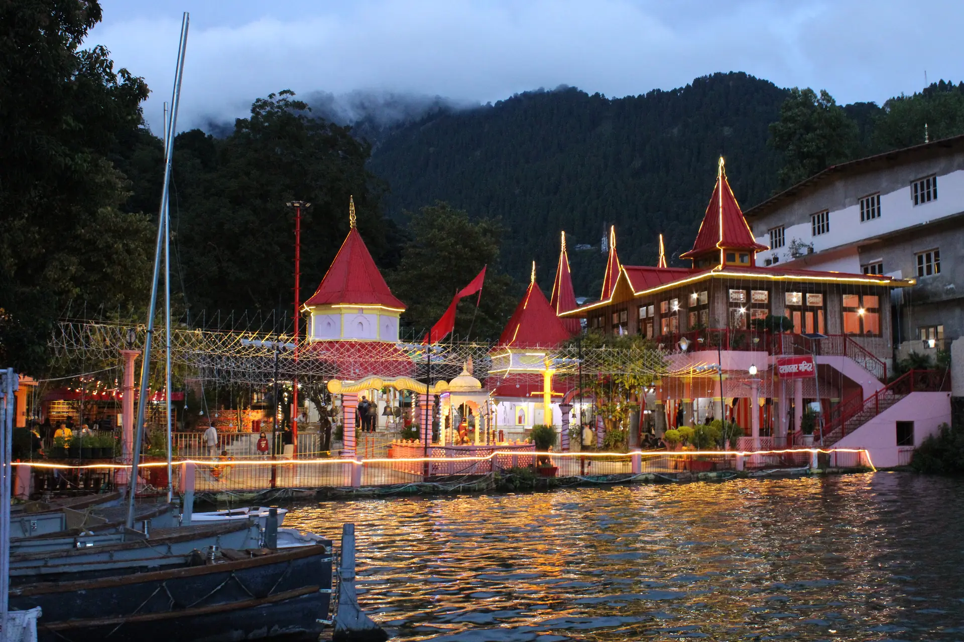 The red-roofed Naina Devi Temple on the shore of Nainital Lake at dusk, lit up, with boats moored in front and forested hills behind