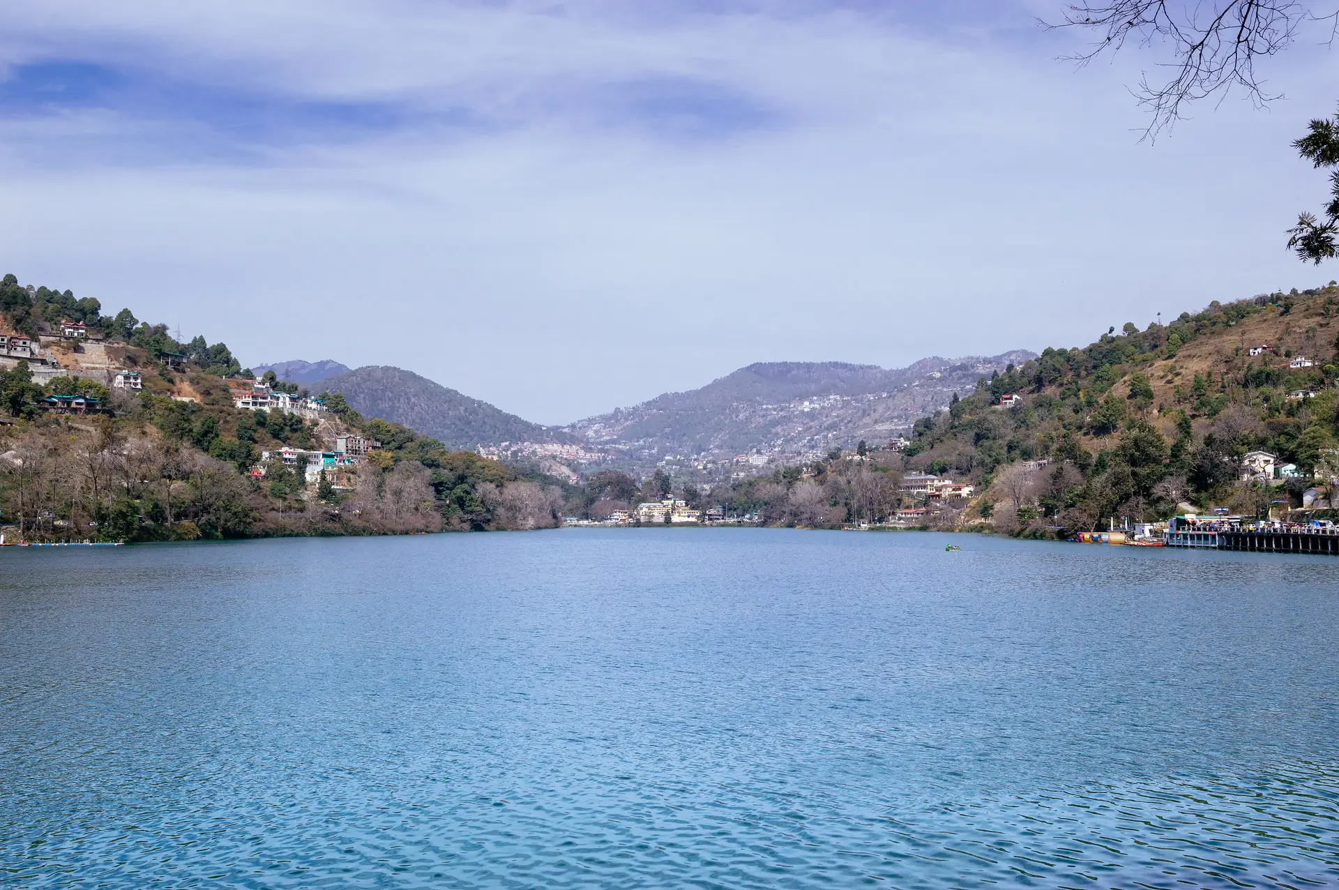 Bhimtal Lake spread out under a blue sky, with houses on the forested slopes on the left and ridges beyond