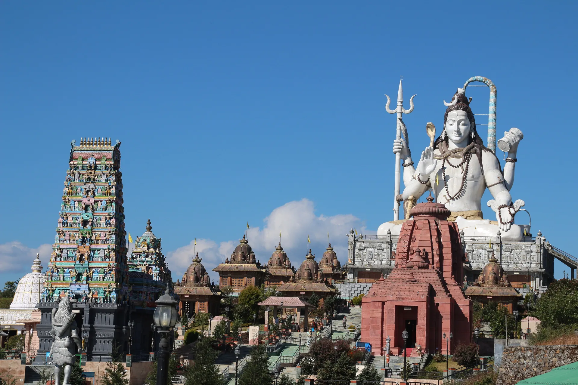 A tall white statue of Shiva with a trident above a cluster of temples and a gateway tower on a hilltop under a blue sky