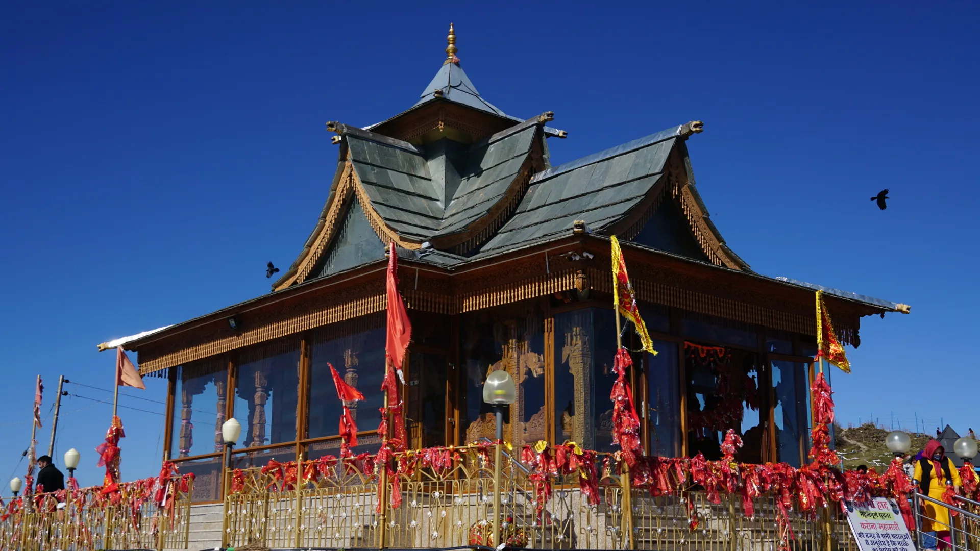 The wooden Hatu Mata Temple with its tiered slate roof and red banners under a blue sky