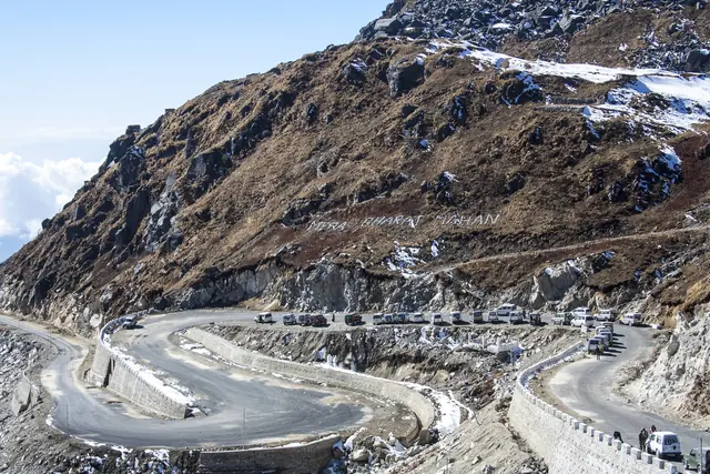 A road climbing in tight hairpins up a steep, bare, snow-streaked mountainside with a line of vehicles on the bends