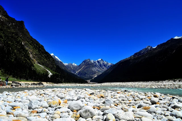 A wide stony river bed in the Yumthang valley between dark mountain slopes, with snow peaks at the head of the valley under a deep blue sky