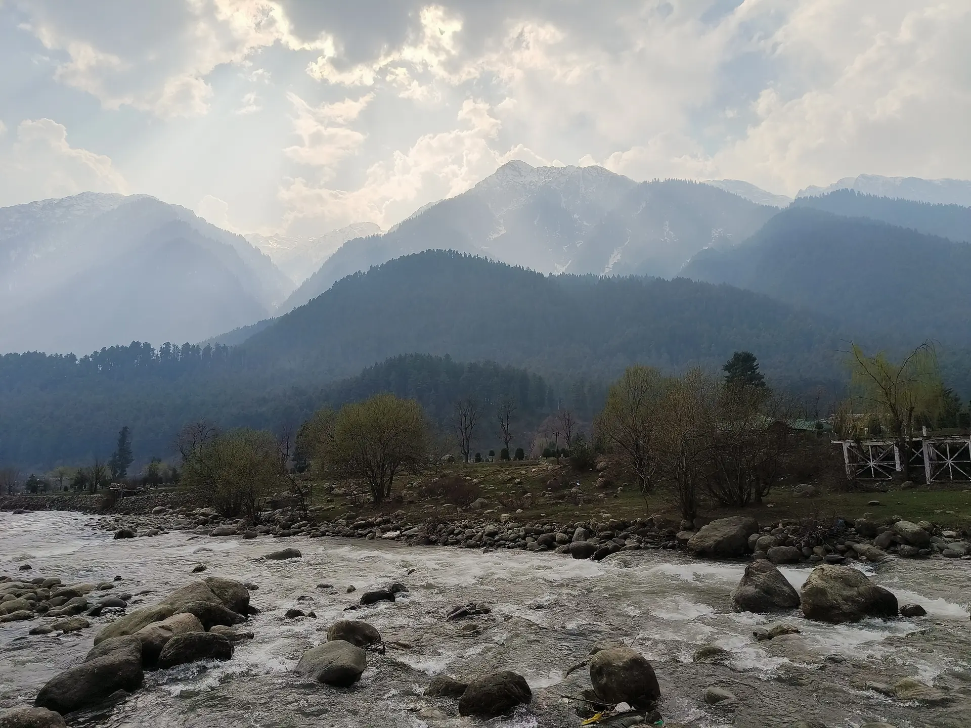 The Lidder river running over boulders in front of tree-covered hills and distant mountains under a cloudy sky