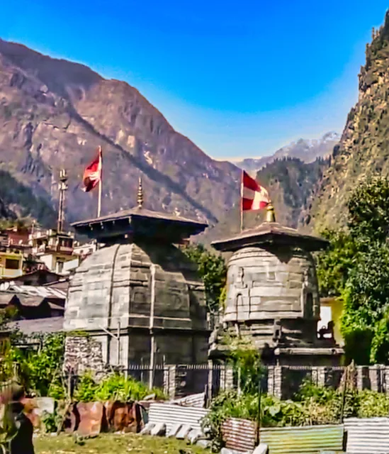 Two small stone temples with tiered towers and red flags at Pandukeshwar, steep green mountains behind