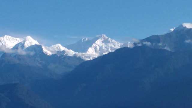A long snow-covered Kanchenjunga ridge in blue morning light above dark forested hills with a few wisps of cloud